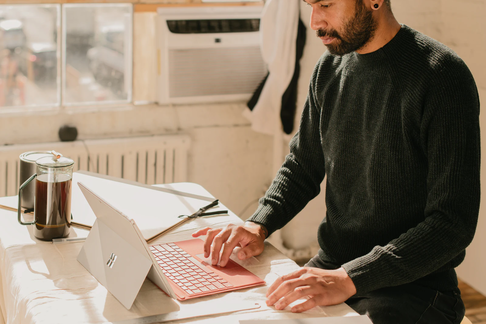 A man typing on a laptop at a bright table, a coffee press beside him