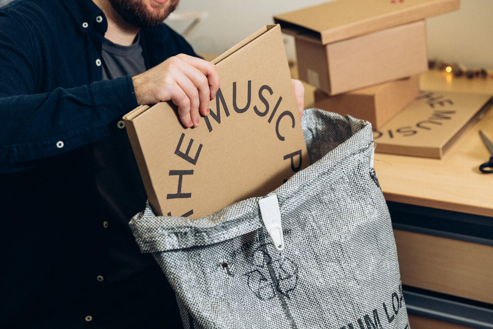 Hands packing a record into a shipping box beside a stack of mailers